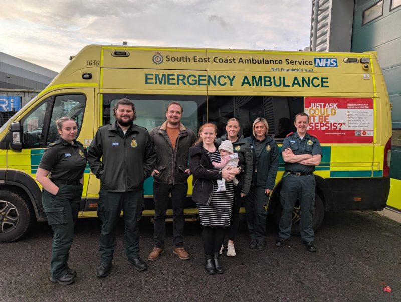 Mother and father holding baby stood with ambulance service clinicians in front of ambulance smiling