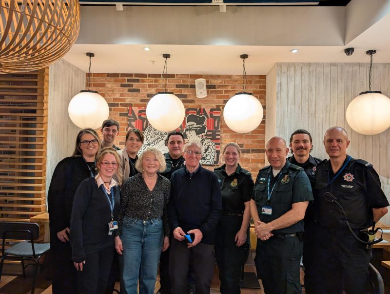 A group of paramedics, fire and rescue staff, passenger operations staff and two members of the public pose together inside a warmly lit restaurant in London Gatwick.