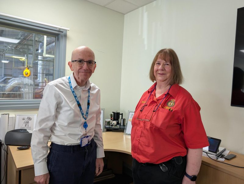SECAmb CEO Simon Weldon in white shirt and blue lanyard stands next to Community First Responder, Sally Holmes, in red uniform shirt in an office with ambulance garage visible through window behind them.