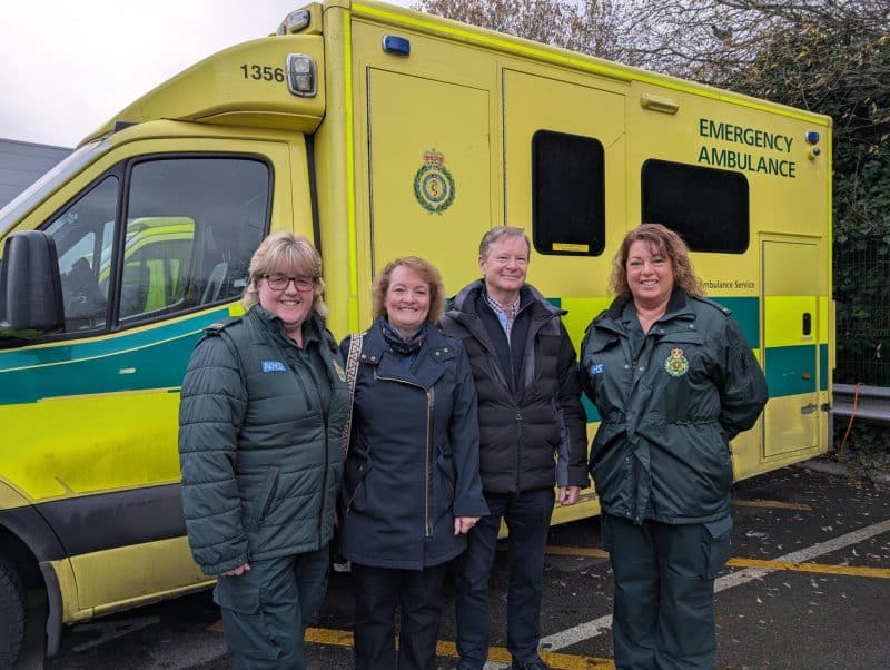 Four people stand in front of a yellow and green emergency ambulance in a car park, with two wearing ambulance service uniforms and two in dark coats.
