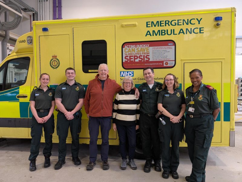 A group of seven people stand in front of a yellow NHS emergency ambulance inside a garage. Two people wear casual clothing, and the others wear green ambulance service uniforms. A sepsis awareness poster and NHS markings are visible on the ambulance.