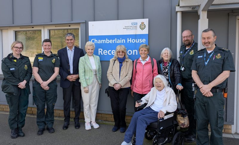 A group of people stand together outside Brighton Make Ready Centre in front of a sign that reads “Chamberlain House Reception,” with some individuals wearing green ambulance service uniforms and others in everyday clothing.