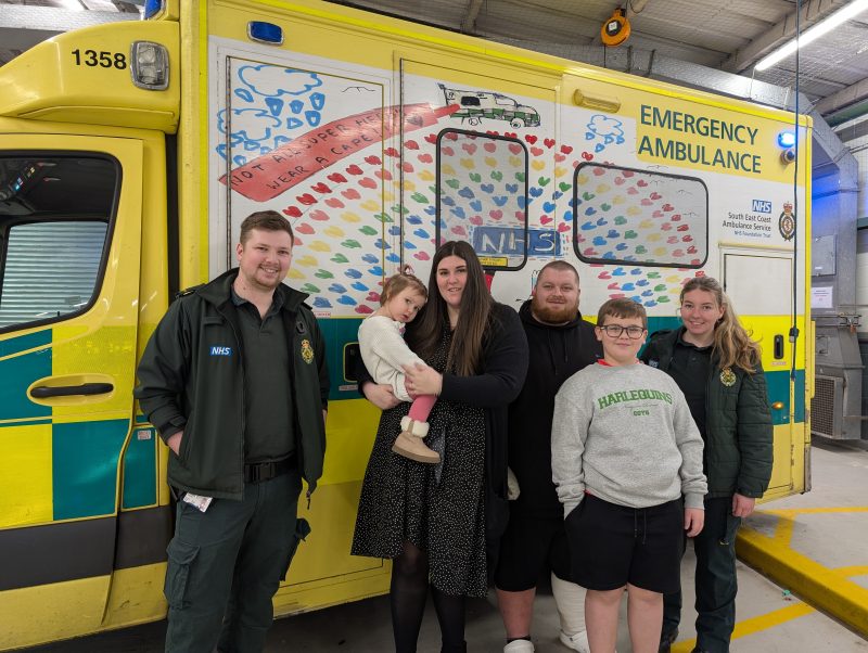 A group of six individuals stands together in front of an emergency ambulance. The ambulance features child‑style artwork on its side panel, including colourful handprints, a rainbow‑like pattern, and small drawings of clouds and an emergency vehicle. Two paramedics on the left are wearing dark green uniforms with NHS markings. The others are dressed in everyday clothing, and one person in the centre is holding a small child.