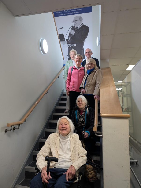 A group of people are positioned on an indoor staircase, with one person seated at the bottom using a mobility aid. The others stand on the steps behind with large black and white image of Douglas Chamberlain on the wall at the top of the stairs.