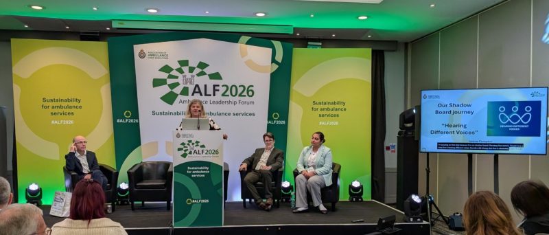 A panel session takes place at the Ambulance Leadership Forum 2026, with one person speaking at a lectern while four others sit on stage. The backdrop features green event branding with the text “Sustainability for ambulance services” and “#ALF2026.” A large screen on the right displays a presentation slide titled “Our Shadow Board journey: Hearing Different Voices.”