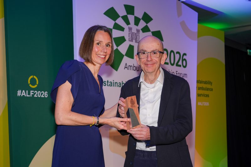 Two people stand on stage at the Ambulance Leadership Forum 2026 as one presents an award to the other, with green and yellow event branding and the hashtag #ALF2026 in the background.