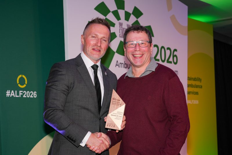 Two people shake hands as one holds an award in front of a backdrop for the Ambulance Leadership Forum 2026, with green event branding and the hashtag #ALF2026.