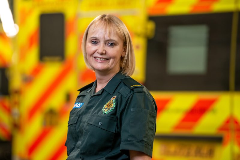 A blonde woman in a paramedic uniform stands before an ambulance