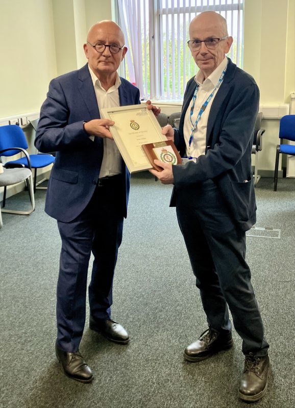 Two people standing in an office setting, jointly holding a framed certificate and a presentation box, with chairs and windows visible in the background.