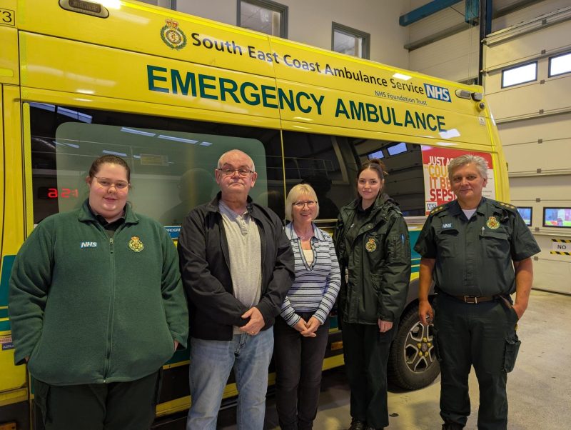 Five adults stand indoors in front of a bright yellow South East Coast Ambulance Service NHS emergency ambulance. They face the camera, wearing a mix of NHS uniforms and casual clothing, inside SECAmb Tangmere Make Ready garage.