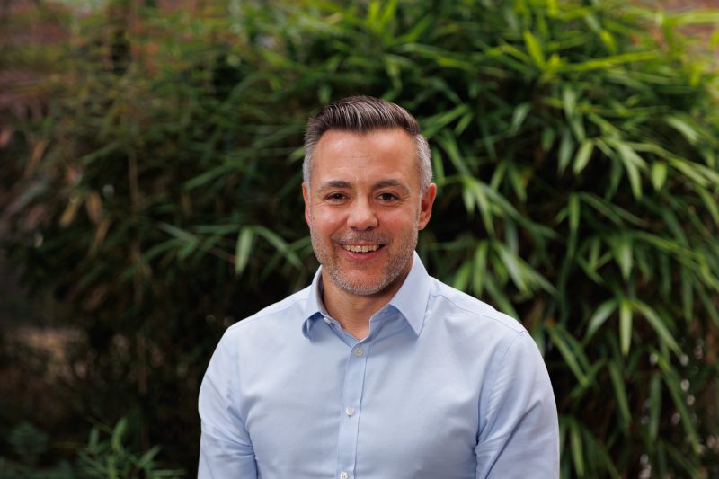 Chief Executive, Simon Ashton, in a light blue shirt standing outdoors, with green foliage in the background.