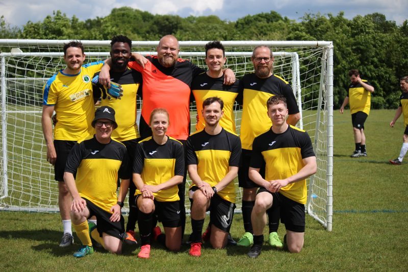 Nine people in yellow football shirts and goalkeeper in orange stand and crouch in two lines in front of football goal