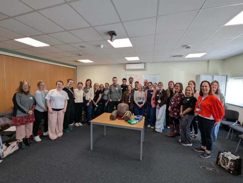 Midwives and ambulance clinicians standing in a meeting room around a table with a maternity training mannequin and supplies.