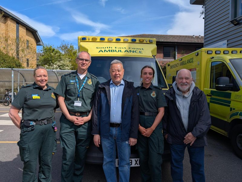 Six adults stand shoulder to shoulder outdoors in front of two yellow emergency ambulances, one marked “South East Coast Ambulance” and “Emergency Ambulance.” Several people wear green emergency service uniforms with visible badges, while others wear casual jackets;