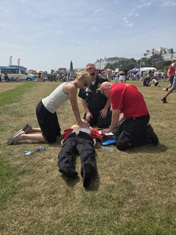 Three people kneel on a grassy field demonstrating CPR on a training mannequin during an outdoor community event - Eastbourne 999 day