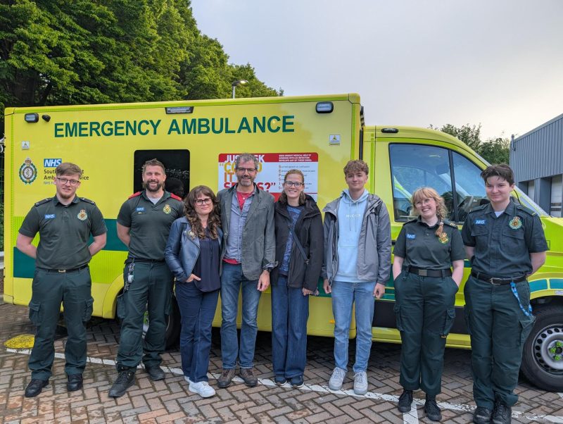 Eight people stand in a row in front of a bright yellow NHS emergency ambulance. Four wear green ambulance uniforms, while four wear casual clothes.