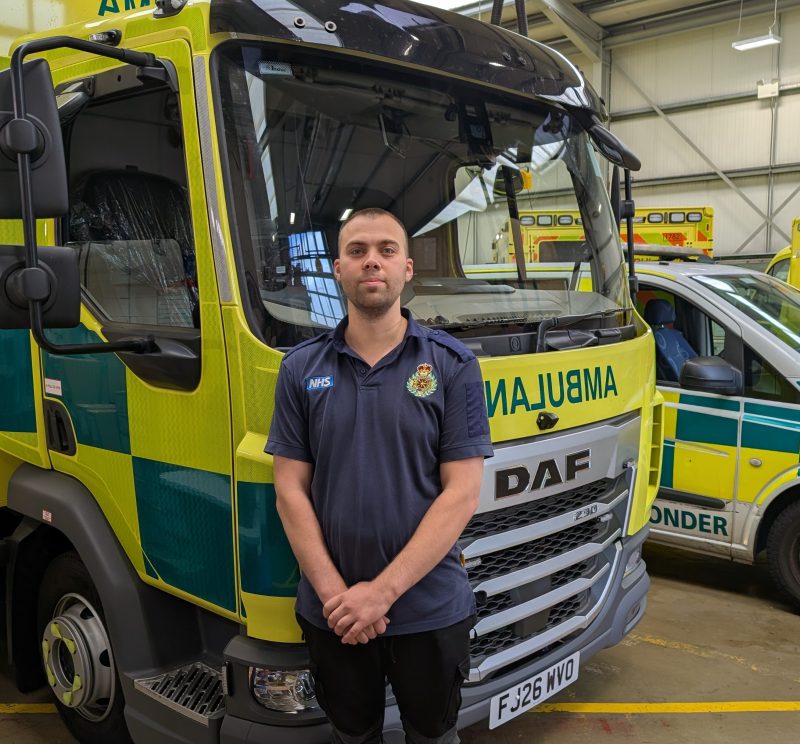 A man with cropped dark hair and a blue polo T-shirt with the SECAmb logo stands in front of an ambulance.