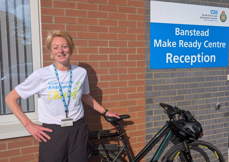 A blonde-haired woman wearing a white T-shirt branded with SECAmb Charity logo stands with her bicycle.