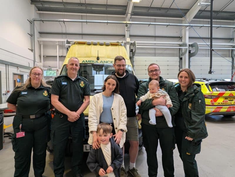 SECAmb ambulance staff and a family with two young children stand together in a garage with ambulances during a reunion at Polegate Make Ready Centre.