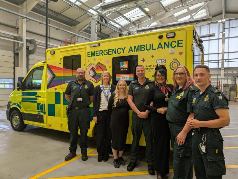 Six ambulance staff stand in a garage beside a yellow emergency ambulance decorated with a rainbow Pride design