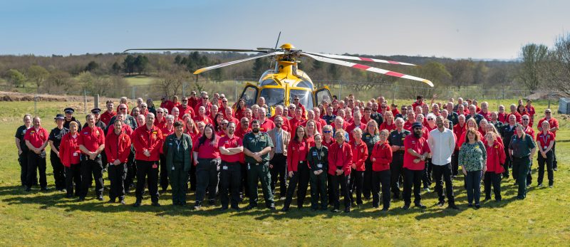 A large group of uniformed volunteers and emergency service personnel stands on a grassy field in front of a bright yellow rescue helicopter