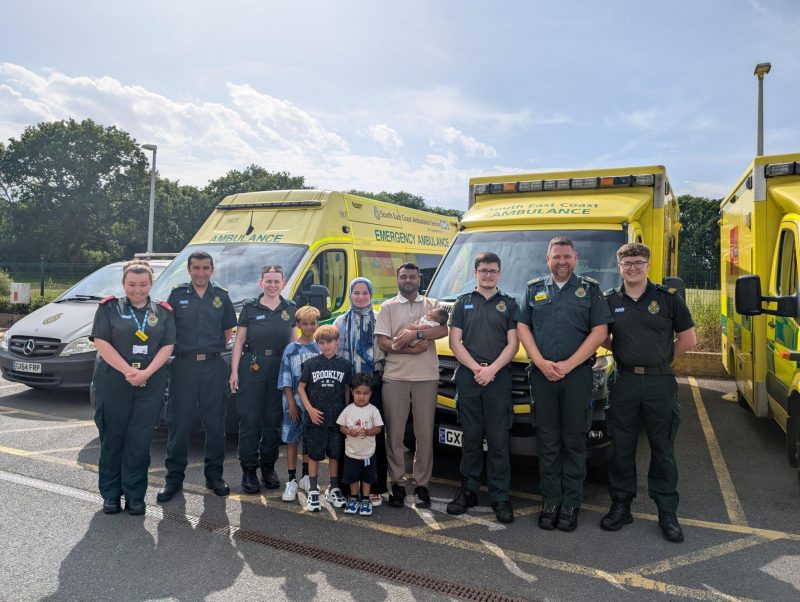 Group photo taken outdoors in an ambulance station car park. Eight ambulance service staff in dark green uniforms stand alongside a family with four children and a baby held in an adult’s arms.