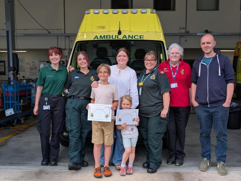 Group photo in front of an ambulance at an ambulance station, with two children holding certificates and ambulance staff, volunteers and family members standing together.