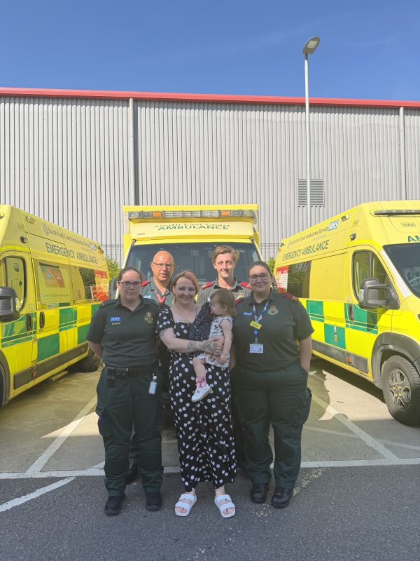 A lady holding her baby standing next to ambulance staff wearing green in front of an ambulance