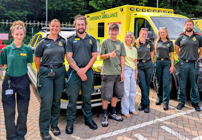 A group of ambulance staff and community members stand in front of two NHS emergency ambulances in an outdoor parking area. One person in the centre gives a thumbs-up gesture.