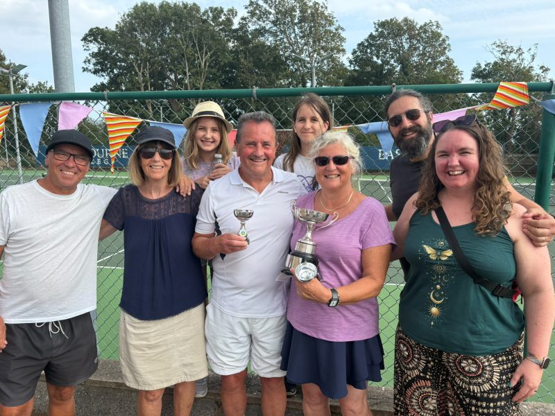 Group standing in front of tennis courts with colourful bunting, with two people holding trophies after a tournament.
