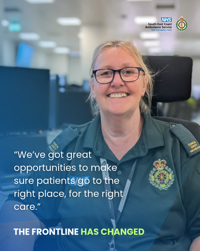 NHS ambulance service staff member seated at a workstation in an office, wearing a green uniform and lanyard.