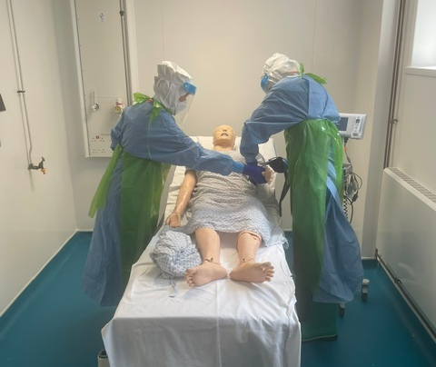 Two people wearing PPE attend to a practice dummy lying on a hospital bed 