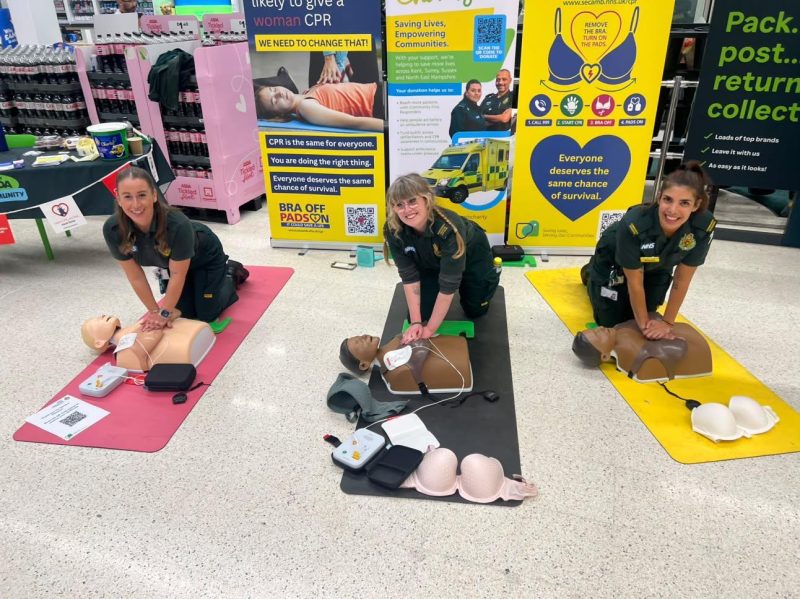 SECAmb colleagues performing CPR on training manikins infront of campaign banners with bra off pads on slogan on